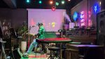 Live music bar scene with a guitarist playing under colorful lights, capturing a quiet pause moment