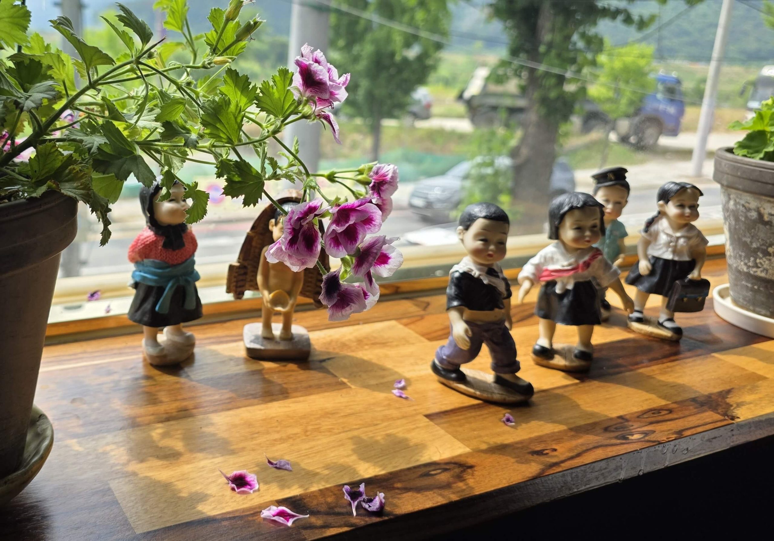 Children figurines by a café window surrounded by flowers, reflecting a quiet moment of growing and belonging