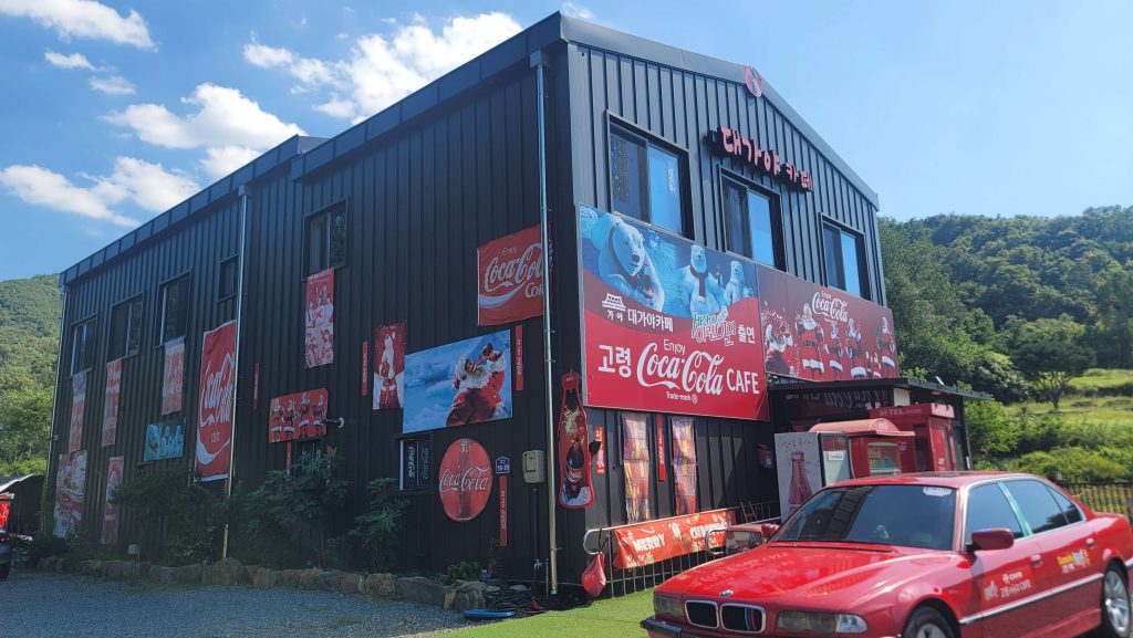 The exterior of a Coca-Cola-themed café in Goryeong surrounded by green hills on a summer afternoon.