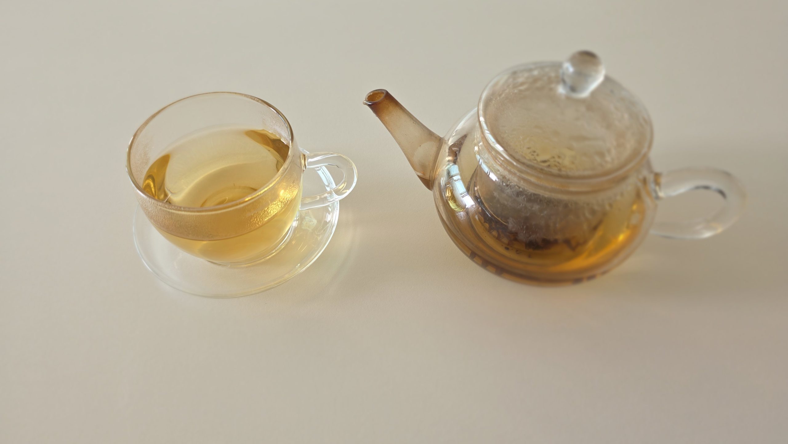 A glass teacup filled with golden black tea beside a glass teapot on a light-colored table.