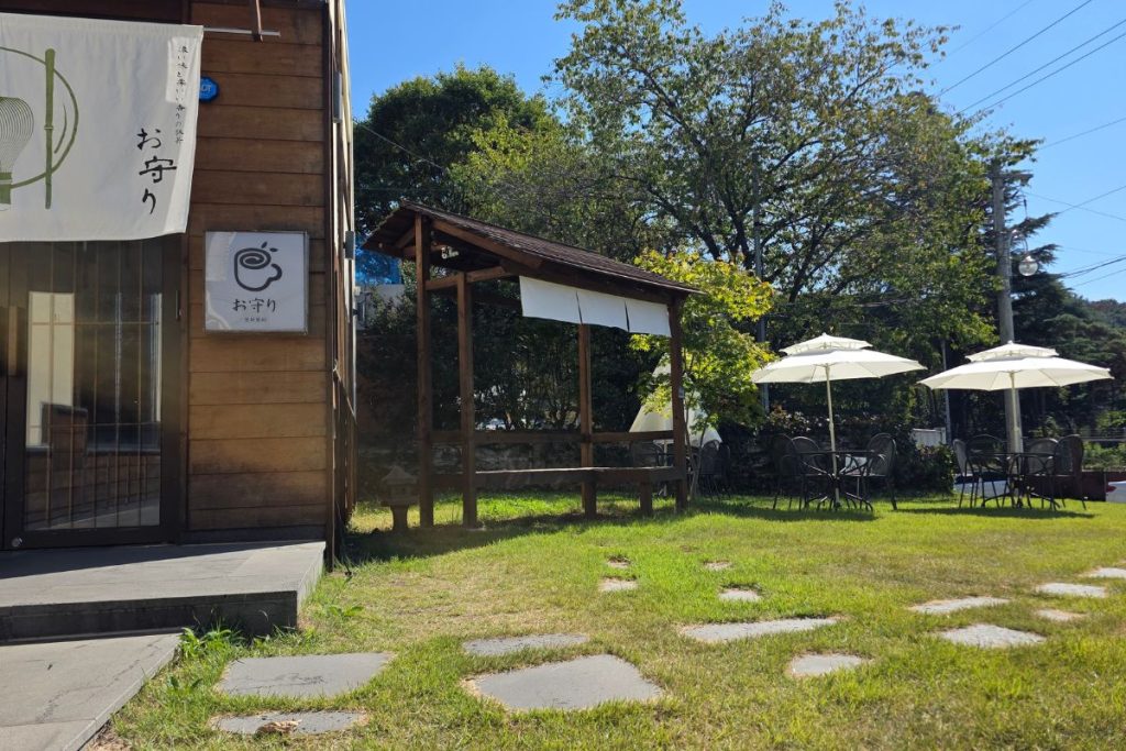 Japanese-style cafe entrance beside a green lawn with stepping stones, outdoor seating, and trees in autumn sunlight.