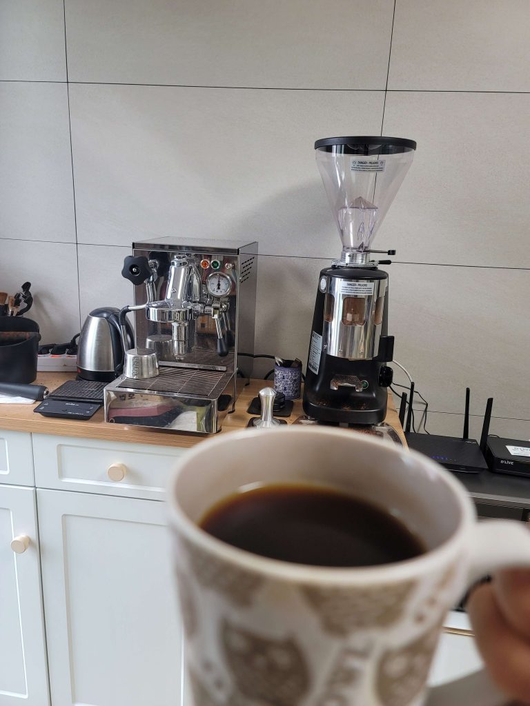 An espresso machine, coffee grinder, and a cup of coffee on a wooden counter in my living room.